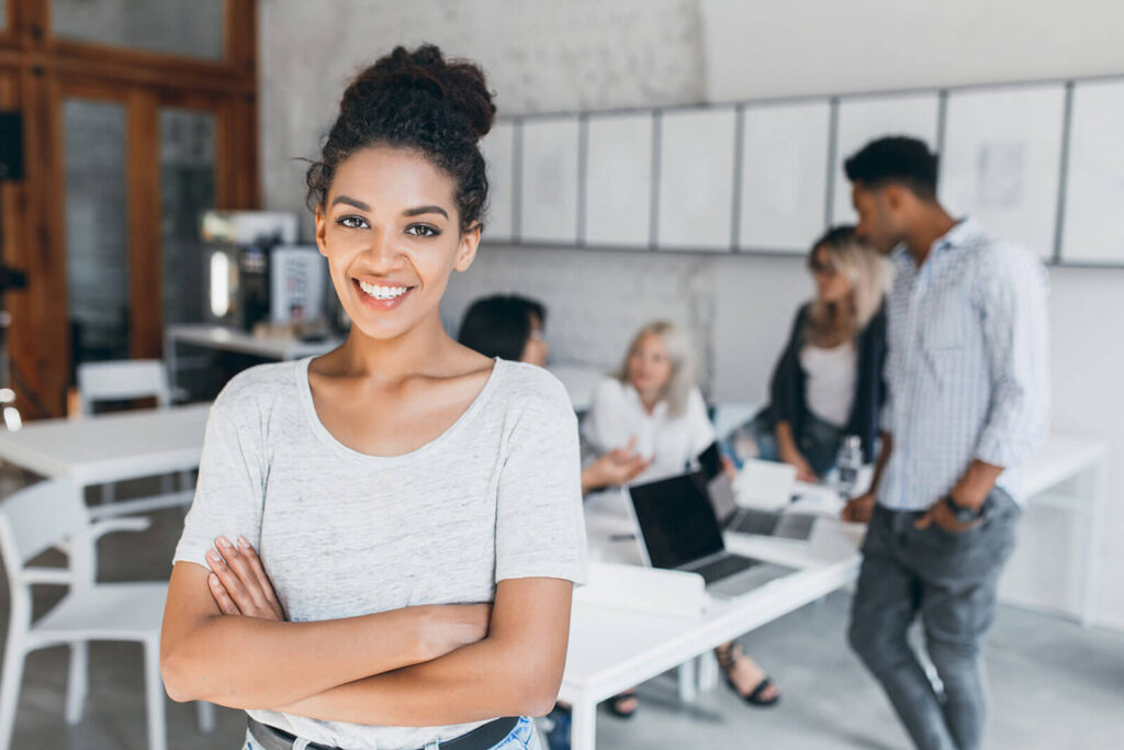 Woman Looking in Camera and Four Others in a Meeting at Workspace Behind